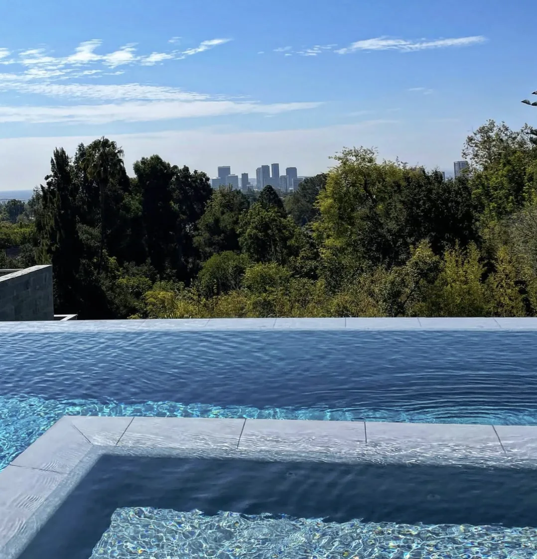 Pool and attached spa with clear blue water overlooking trees and a city skyline